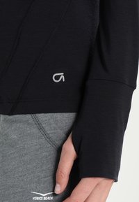 Svart långärmad topp med tumhål, med texturerade sömmar och en liten grå logotyp. Matchas med grå sportbyxor.