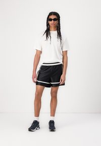 T-shirt blanc, shorts de sport noirs avec des bordures blanches et baskets noires. Le modèle a de longs cheveux tressés et des lunettes de soleil. Fond uni.