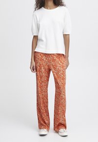 Bílý krátký rukáv top spárovaný s širokými kalhotami s oranžovým květinovým vzorem. Materiály vypadají měkce; kalhoty mají kombinaci oranžových a béžových akcentů.