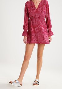 Blumenmusterkleid in lebhaftem Pink mit langen Ärmeln, einem V-Ausschnitt und einem elastischen Bund, kombiniert mit silbernen Slipper-Sandalen.