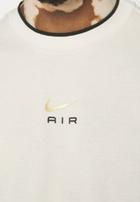 Jasno beżowy sweatshirt z okrągłym dekoltem i czarnym wykończeniem. Posiada złoty znak Nike oraz słowo "AIR" w czerni poniżej.