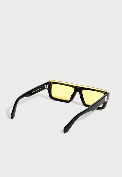 Lunettes de soleil rectangulaires noires avec verres teintés jaunes et branches épaisses, vues en angle arrière sur fond blanc.