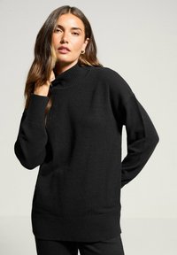Schwarzer Rollkragenpullover mit gerippten Bündchen und Saum, lockere Passform und weicher Textur, getragen über schwarzen Hosen.