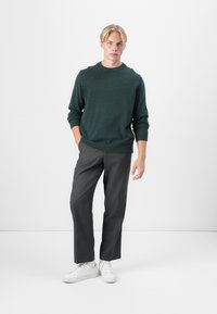 Pulover tricotat de culoare verde închis cu guler rotund, asociat cu pantaloni gri adaptați și adidași albi. Modelul stă pe un fundal neutru.