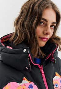 Padded bunda s čiernym povrchom, ružovými akcentmi a farebnými grafickými vzormi. Má vysoký golier a viditeľný zips na zapínanie.