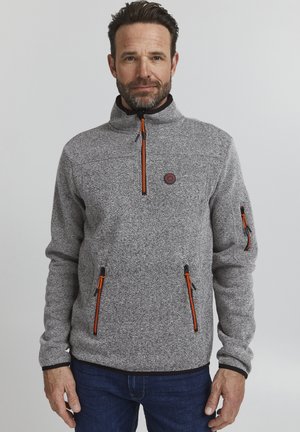 Grå fleece-genser med høy krage, med glidelåsdrager i svart og oransje og to framlommer med glidelås. Teksturert stoff med logo-detalj.