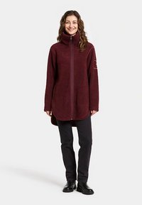 Veste en polaire bordeaux avec col montant, fermeture éclair intégrale à l'avant, manches longues et ourlet arrondi. Texture lisse ; logo sur la manche droite.