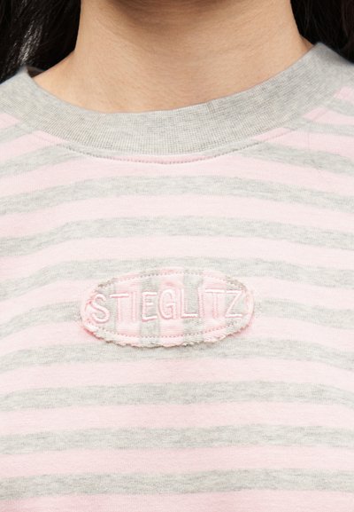 Κοντινό πλάνο του λαιμού και του πάνω μέρους του στήθους ενός ατόμου που φοράει μια ροζ και γκρι ριγέ μπλούζα με ραμμένη οβάλ ετικέτα που γράφει «STIEGLITZ».