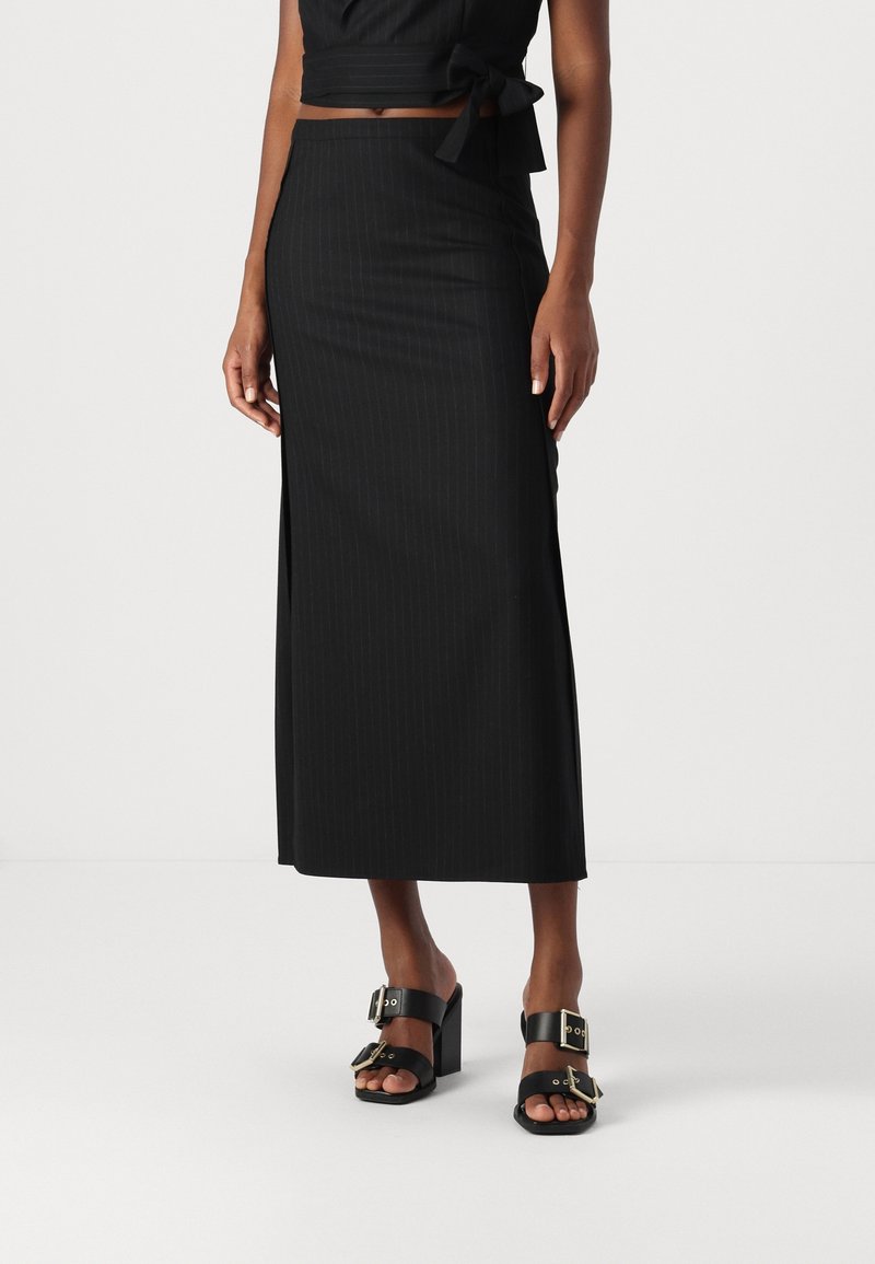 Femme portant une jupe midi noire taille haute avec de fines rayures discrètes et des sandales à talons noires avec des boucles dorées.