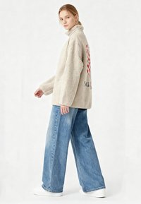 Giovane donna che indossa una giacca in pile beige con un disegno a cuore rosso sulla schiena, jeans blu a gamba larga e sneakers bianche, in piedi davanti a uno sfondo bianco.