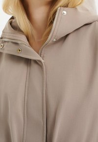 Béžová bunda s kapucí, vybavená zipem a cvočky, vyrobená z hladké látky s minimalistickým designem a bez viditelných vzorů.