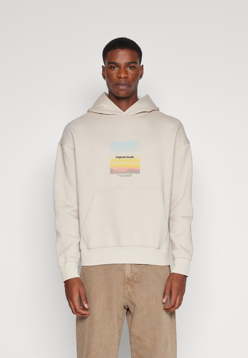 Lichtbeige hoodie met een voorzak en een trekkoordkap. Heeft een rechthoekige graphic in pastelkleuren, gelabeld "Originals Studio."