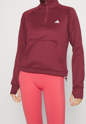 Bordowy pulower z półzamkiem, przednią kieszenią i białym logo, noszony z koralowymi legginsami z wysokim stanem na stojącej osobie.