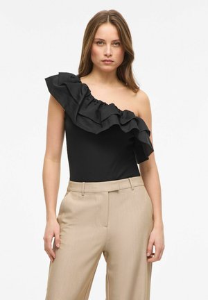 Jeune femme portant un pantalon beige et un top noir à une épaule avec un grand volant sur l'épaule gauche, debout devant un fond blanc.