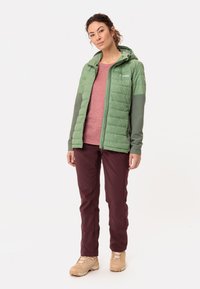 Groene geïsoleerde jas met grijze panelen, bordeauxrode broek en beige laarzen. De outfit bevat een capuchon en een textuurstofafwerking.