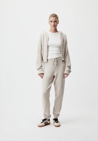 Felpa con zip beige in maglia e pantaloni jogger coordinati, con polsi a coste e vita con coulisse. Il modello indossa sneakers nere e bianche.