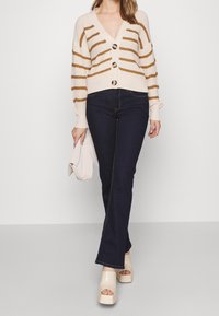 Nainen, jolla on yllään vaaleanbeige raidallinen neuletakki suurilla napeilla, tummansiniset farkut, vaaleanbeige korkkipohjaiset sandaalit ja hän pitää kevyttä vaaleanbeigeä clutch-laukkua.