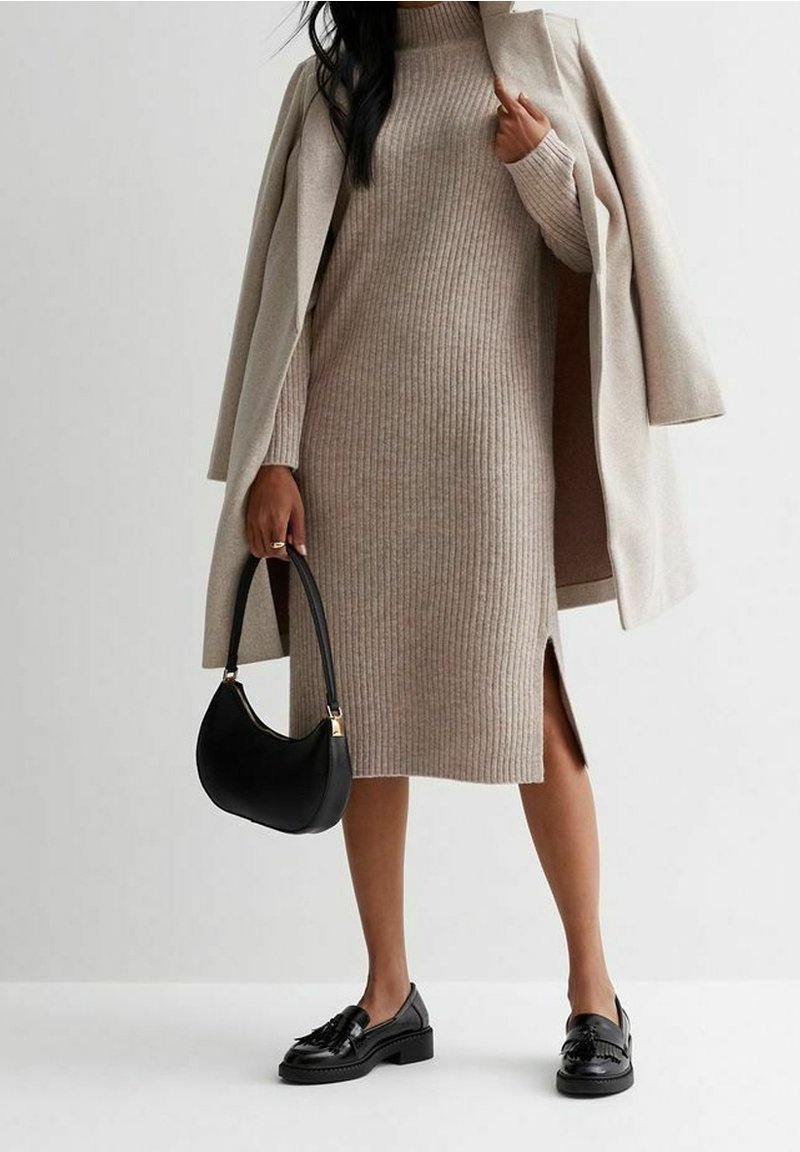 Femme vêtue d'une robe en maille côtelée beige et d'un manteau, tenant un sac à main noir, portant des mocassins noirs à pompons sur un fond uni.