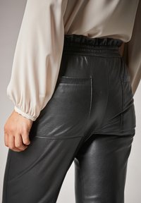 Pantalons en faux cuir noir avec une texture brillante, présentant une taille smockée et deux poches arrière, associés à un chemisier couleur crème.
