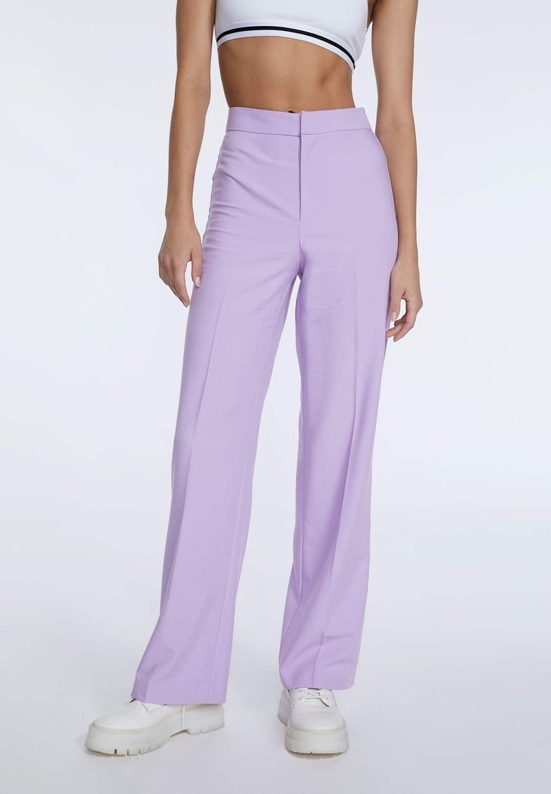 Femme portant un pantalon large taille haute lavande avec un pli devant, des baskets à plateforme blanches et un crop top blanc à rayures noires.
