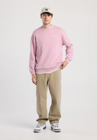 Felpa rosa con scollatura rotonda e testo ricamato, abbinata a pantaloni beige. Modello che indossa un berretto a righe e sneakers. Sfondo semplice.