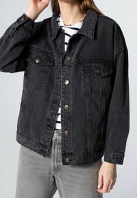 Giacca in denim nero con vestibilità ampia, chiusura con bottoni, due tasche sul petto e dettagli di cucitura a vista. Indossata sopra una camicia a righe bianca e nere.