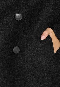 Primer plano de un abrigo de lana negro, con una superficie texturizada, dos botones redondos y un bolsillo lateral donde se ve una mano sosteniéndolo.