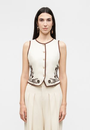 Donna che indossa un gilet crema senza maniche con rifinitura marrone e ricamo floreale, abbinato a pantaloni crema plissettati a vita alta, sfondo neutro.