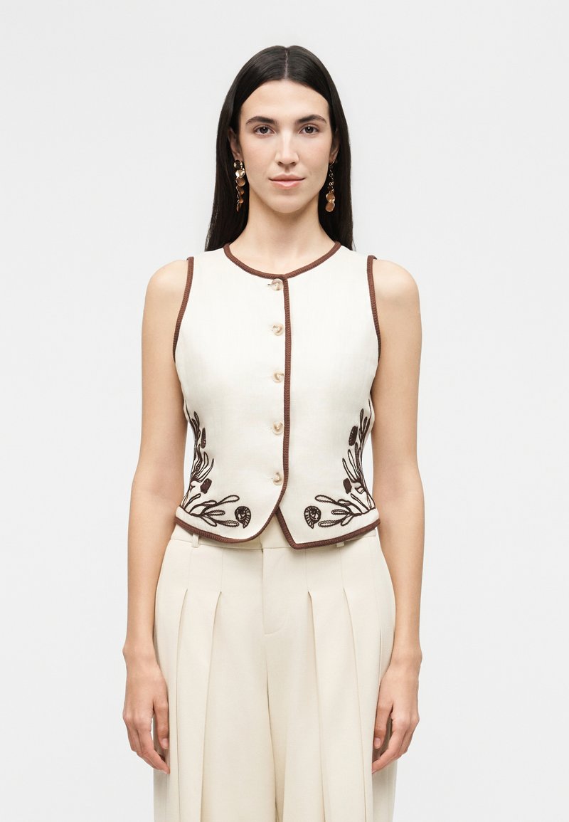 Γυναίκα που φοράει αμάνικο εκρού γιλέκο με καφέ διακοσμητική λωρίδα και κεντήματα με λουλούδια, συνδυασμένο με εκρού πλισέ παντελόνια με ψηλή μέση, ουδέτερο φόντο.