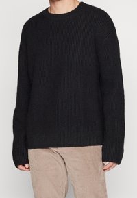 Svart stickad tröja med rund halsringning, sänkta axlar och långa ärmar. Ribbad struktur genom hela tröjan. Matchas med beige kordflorsbyxor.