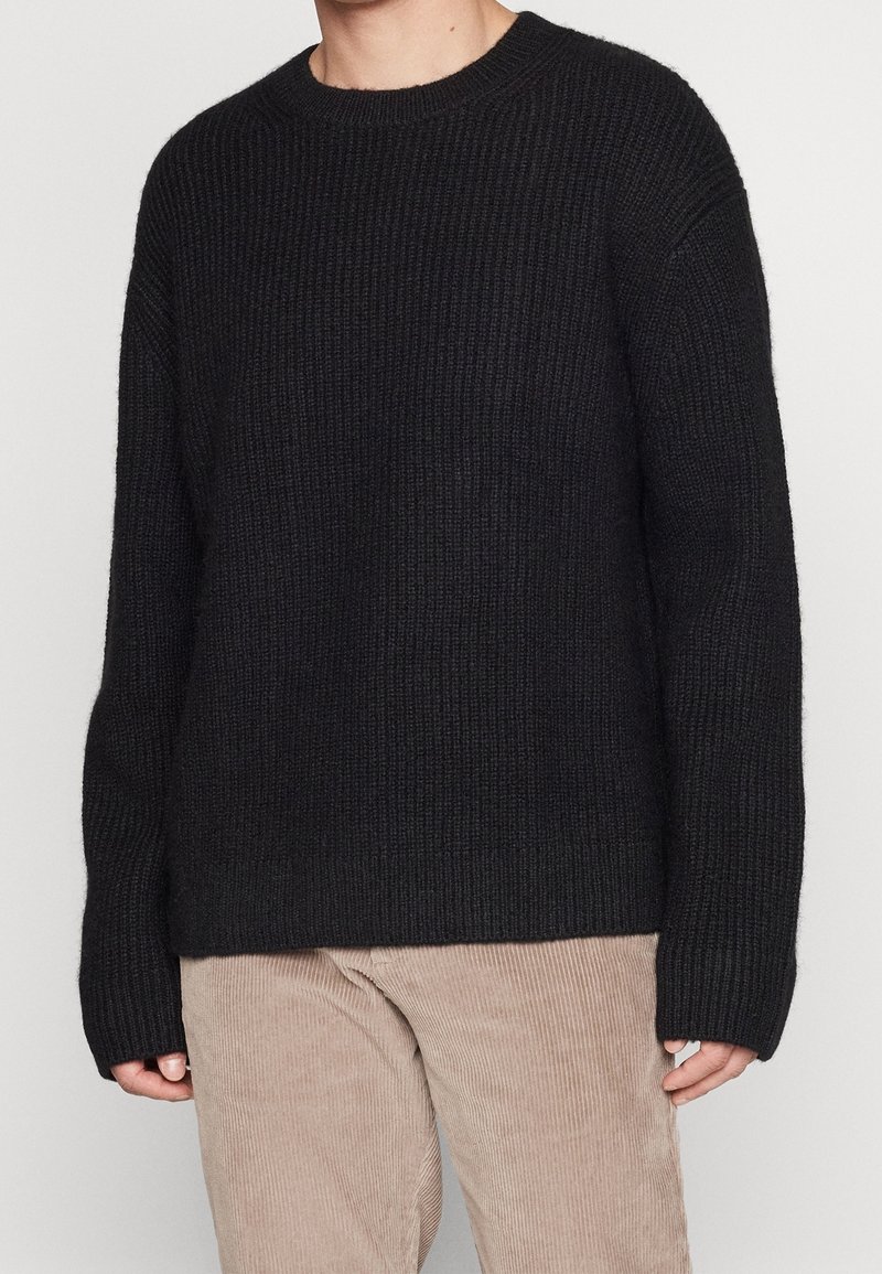 Svart stickad tröja med rund halsringning, sänkta axlar och långa ärmar. Ribbad struktur genom hela tröjan. Matchas med beige kordflorsbyxor.