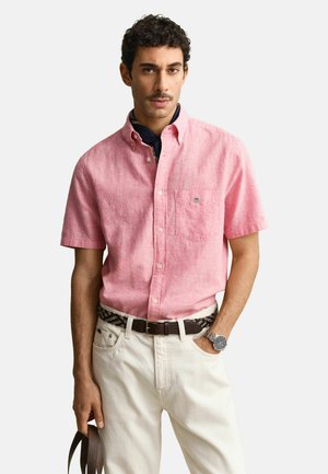 Uomo che indossa una camicia rosa a maniche corte con bottoni, pantaloni bianchi, cintura marrone, orologio da polso e tiene una tracolla marrone.