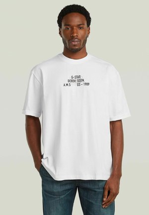 Homme portant un t-shirt blanc à manches courtes avec texte noir et un jean bleu, debout avec une main dans la poche devant un fond uni.