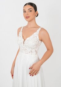 Abito bianco con un corpetto in pizzo decorato da motivi floreali, scollatura profonda e spalline larghe; gonna fluida realizzata in tessuto leggero.