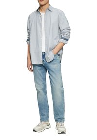 Lichtblauwe, button-up shirt met opgerolde mouwen; gecombineerd met lichtgekleurde jeans en beige sneakers met groene en zwarte accenten.