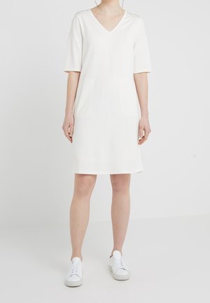 Biała sukienka z krótkim rękawem i dekoltem w serek z dwiema przednimi kieszeniami. Tkanina wydaje się miękka i gładka. Stylizowana z białymi trampkami dla kontrastu.