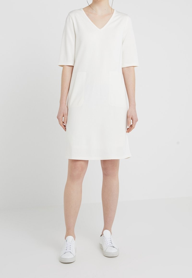 Vestido blanco de manga corta y escote en V con dos bolsillos frontales. La tela parece suave y lisa. Combinado con zapatillas blancas para un contraste.