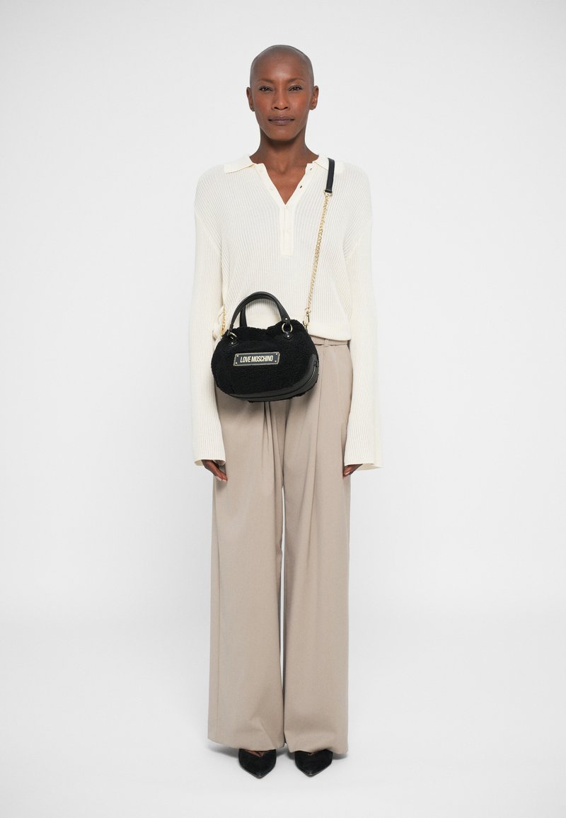 Woman wearing a white knit sweater and beige wide-leg pants, holding a black Love Moschino handbag with a gold chain strap.