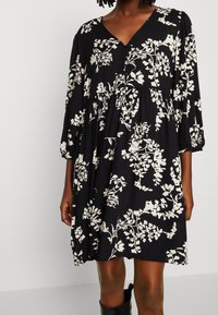 Robe noire avec motif floral blanc, décolleté en V, manches trois-quarts, et tissu froncé pour un ajustement ample, faite en matériau léger.