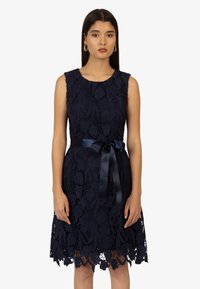 Robe en dentelle bleu marine avec motifs floraux, au design sans manches, décolleté rond, et une ceinture en satin nouée à la taille.