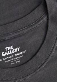 Κοντινό πλάνο στο γιακά σκούρου γκρι T-shirt Jack & Jones Originals με εκτυπωμένη ετικέτα που αναγράφει "The Gallery", κατασκευασμένο στο Μπαγκλαντές, μέγεθος Large.