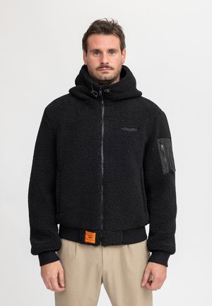 Mann trägt eine schwarze, strukturierte Jacke mit Reißverschluss und Kapuze sowie beige Hose, steht vor einem schlichten hellen Hintergrund.