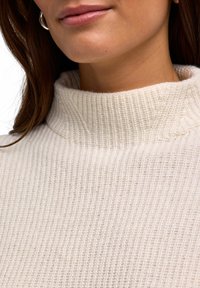 Maglione a collo alto color crema con texture rigata e collo alto. La vista ravvicinata evidenzia i dettagli del tessuto e la forma del scollo.