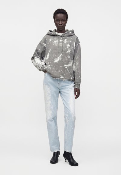 Μαύρο άτομο φορά γκρι φούτερ με tie-dye, ανοιχτό μπλε ξεβαμμένα τζιν και μαύρα μποτάκια μέχρι τον αστράγαλο, στέκεται μπροστά σε απλό ανοιχτό φόντο.