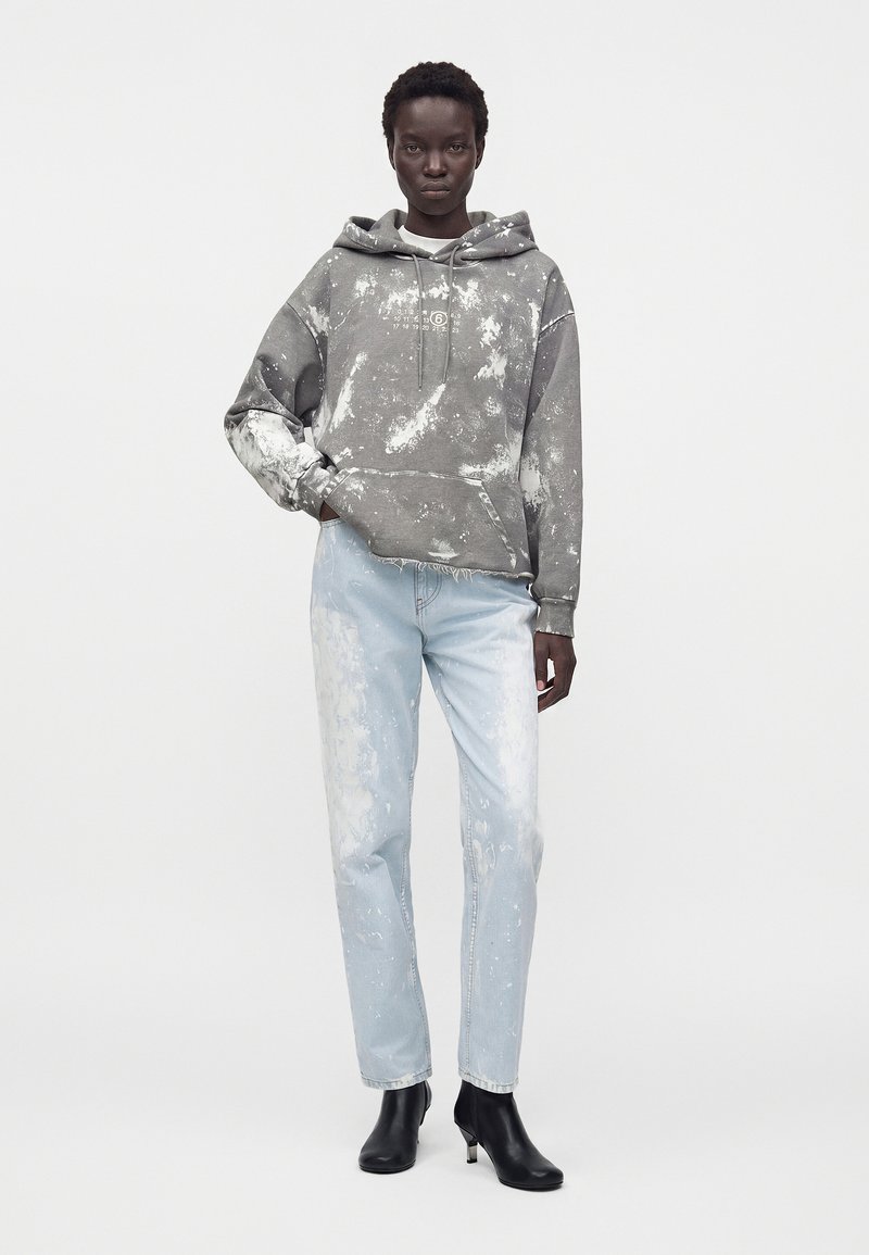 Black person wearing gray tie-dye hoodie, light blue bleached jeans, and black ankle boots, standing against a plain light background.