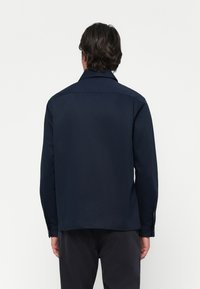 Man wearing a dark navy long-sleeve shirt viewed from the back against a plain white background.