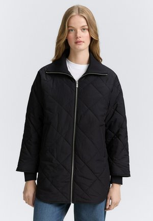 Młoda kobieta o długich blond włosach, ubrana w czarną pikowaną kurtkę na zamek, na białą koszulę i niebieskie dżinsy, stojąca na jednolitym tle.