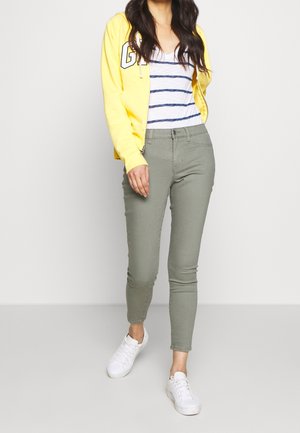 Femme portant un sweat à capuche jaune, un haut à rayures blanches et bleues, un pantalon vert olive et des baskets blanches, debout devant un fond uni.