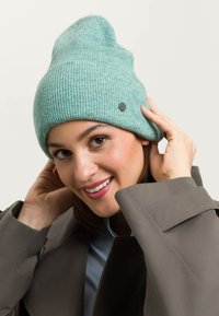 O căciulă tricotată de culoare verde-albastră, cu o textură canalată și un mic accent cu logo negru, potrivită strâns și ușor lăsată în partea de sus.