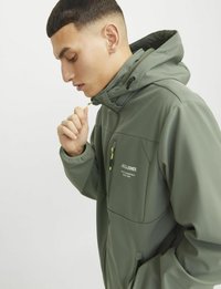 Groene softshell jas met capuchon, ritssluiting aan de voorkant en twee zijzakken. Voorzien van een zwart logo en details in gele rits. Glad materiaal.